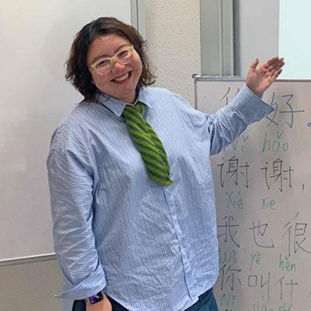 Sun Xiangxue mit Brille, blauem Hemd und grüner Krawatte steht im Klassenraum und zeigt auf eine Tafel mit chinesischen Schriftzeichen